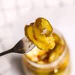 A fork holds up a slice of sweet and spicy pickle above an open jar of pickles, with a blurred checkered cloth in the background.