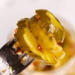 A close-up of sliced pickled jalapeños on a fork, with a jar of Sweet and Spicy Pickles blurred in the background.