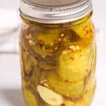 A glass jar filled with sweet and spicy pickles and aromatic spices, sealed with a metal lid, sits on a white surface.