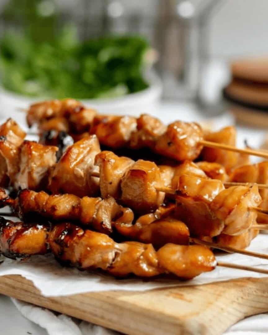 Several skewers of teriyaki chicken on a stick are placed on a wooden board, with a leafy green garnish blurred in the background.
