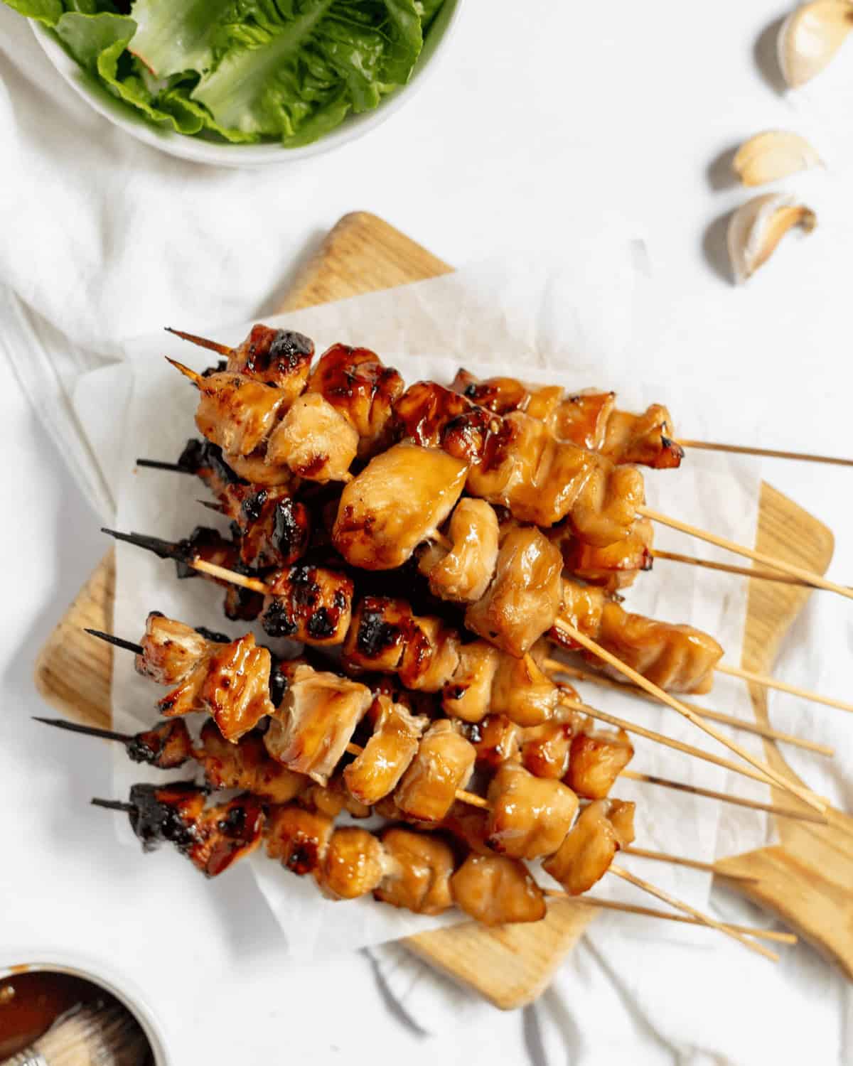 Four baked Teriyaki Chicken on a Stick kebabs are placed on a wooden cutting board with parchment paper. A bowl of greens, garlic cloves, and a condiment dish are visible in the background.