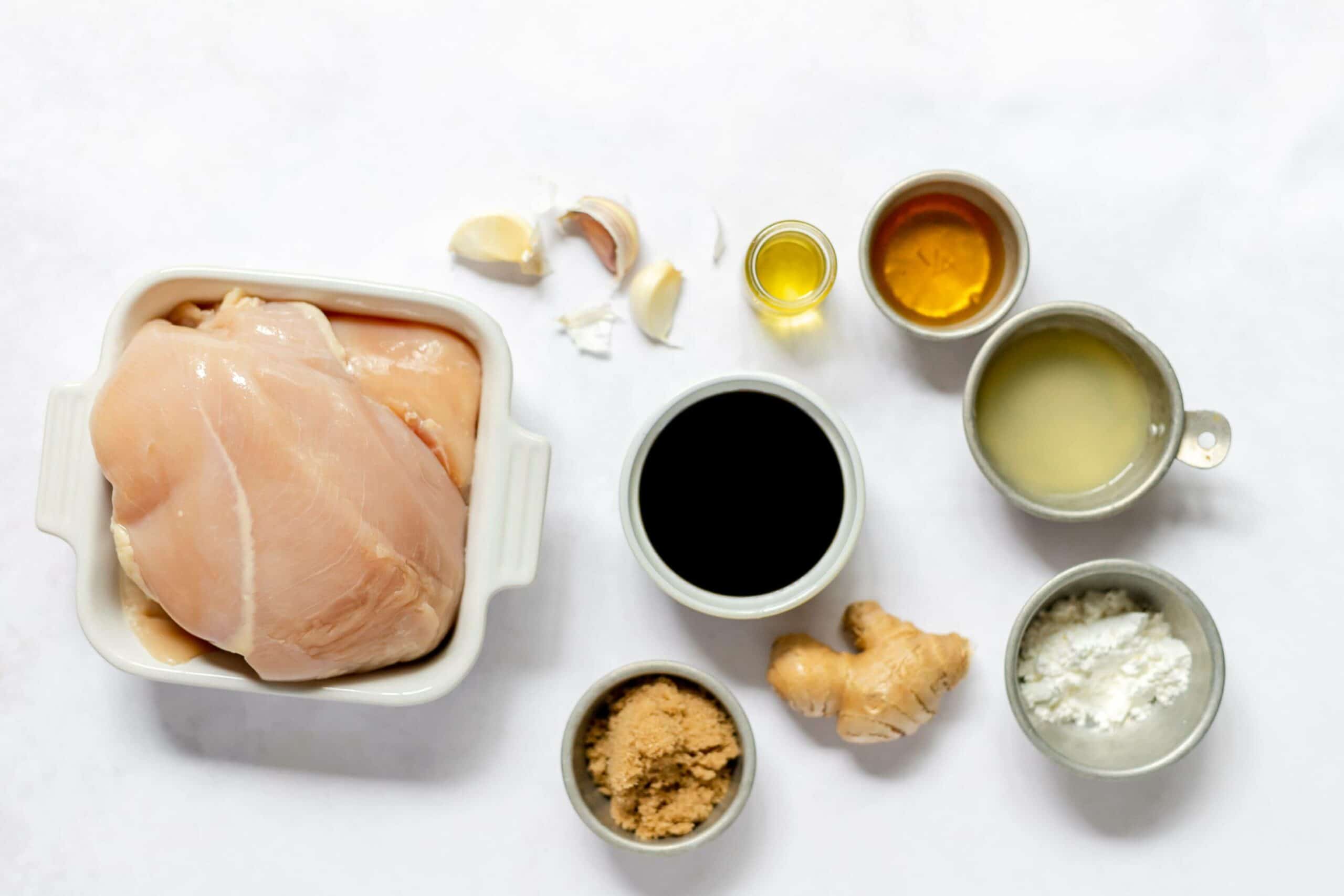 Raw chicken breasts in a white dish surrounded by small containers of garlic cloves, olive oil, honey, lemon juice, soy sauce, brown sugar, ginger, and cornstarch on a white background—perfect ingredients for making Teriyaki Chicken on a Stick.