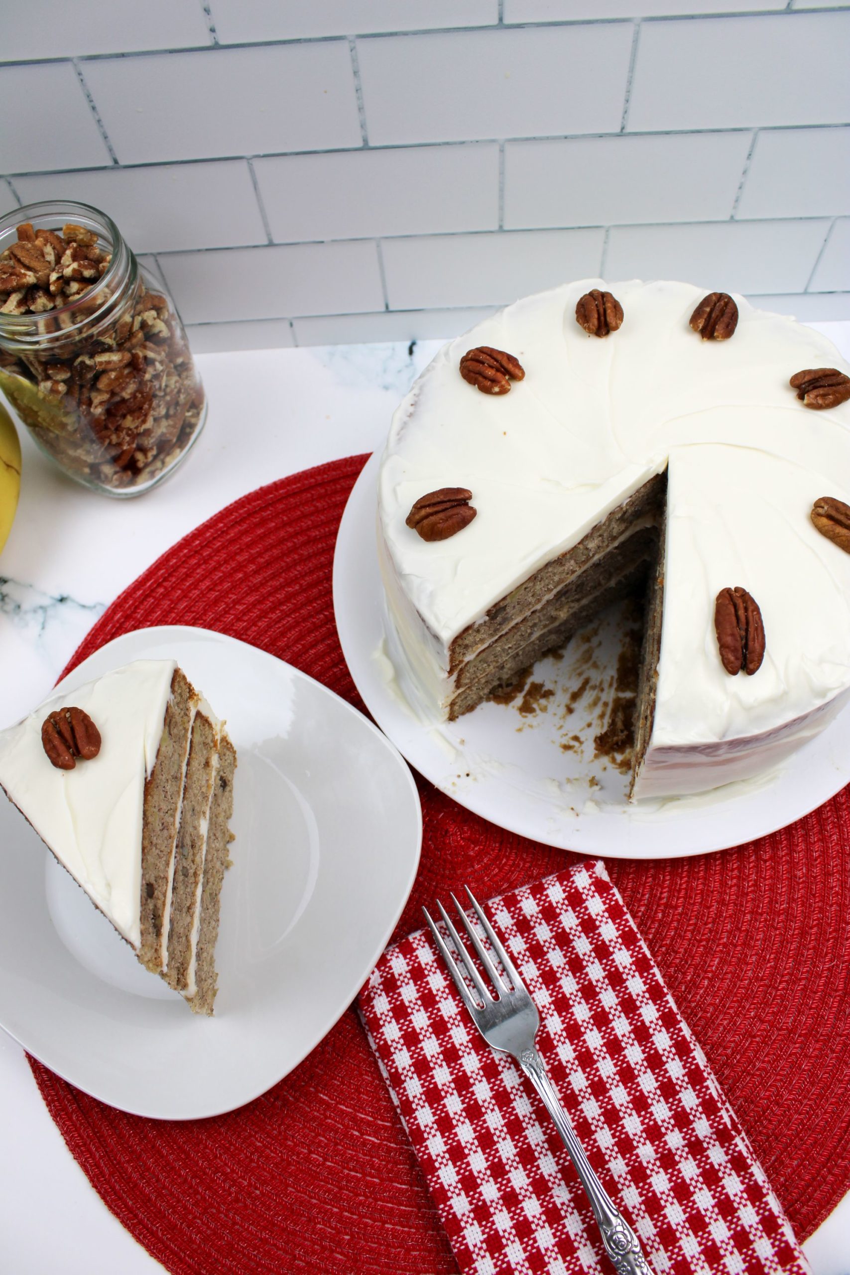 A sliced hummingbird cake with cream cheese frosting and whole pecans on top, displayed on a white plate with a red woven placemat.