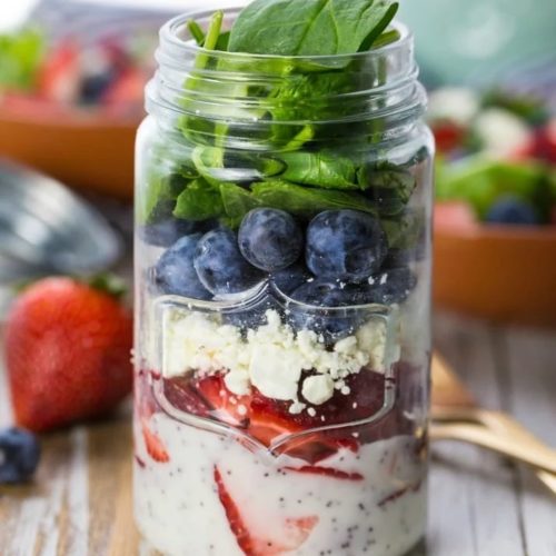 A mason jar filled with strawberries, feta cheese, blueberries, kale, and poppyseeds