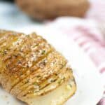 An Air Fryer Hasselback Potato, sliced and baked to perfection, is topped with herbs and coarse salt on a white plate, with a whole potato and striped cloth in the background.