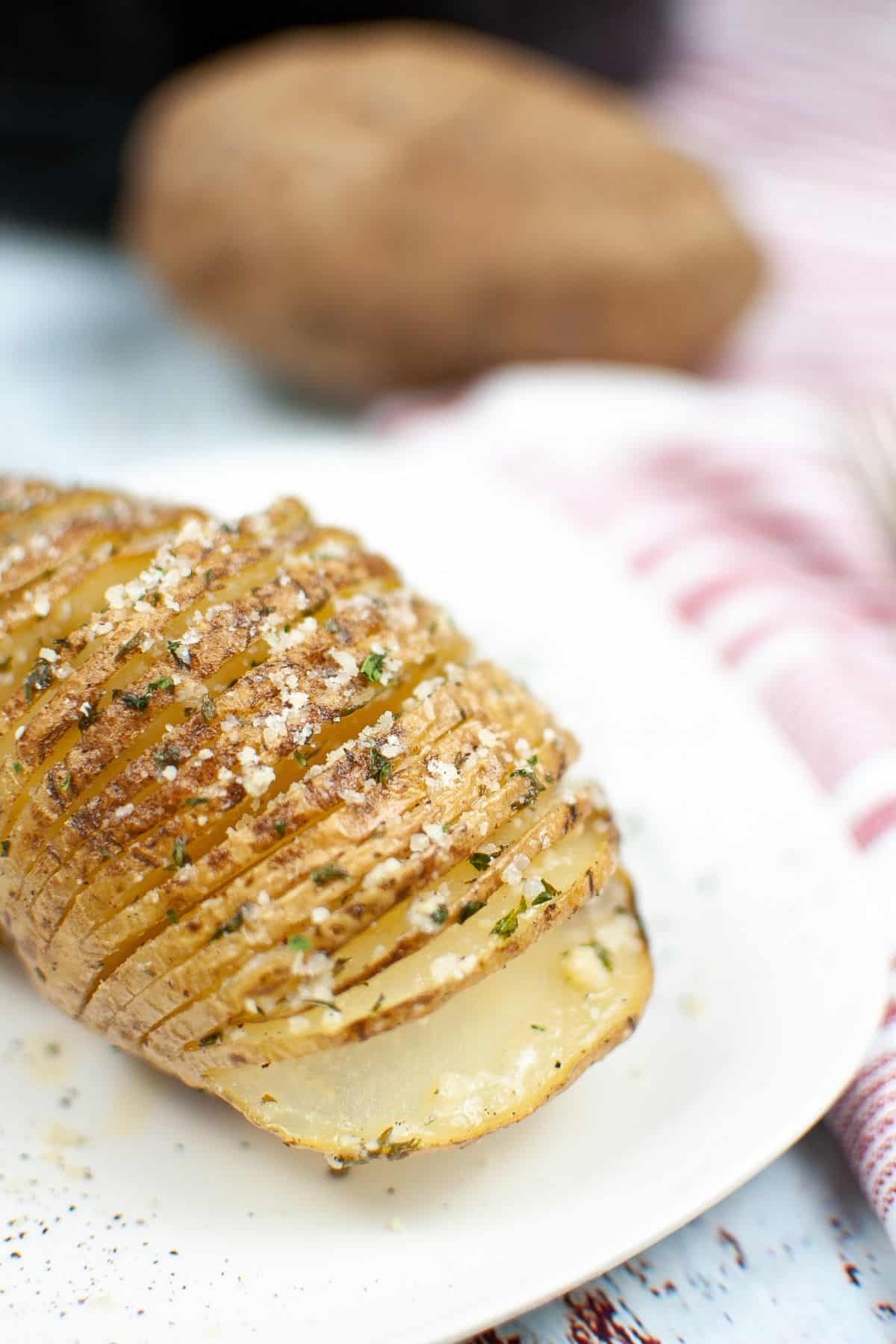 An Air Fryer Hasselback Potato, sliced and baked to perfection, is topped with herbs and coarse salt on a white plate, with a whole potato and striped cloth in the background.