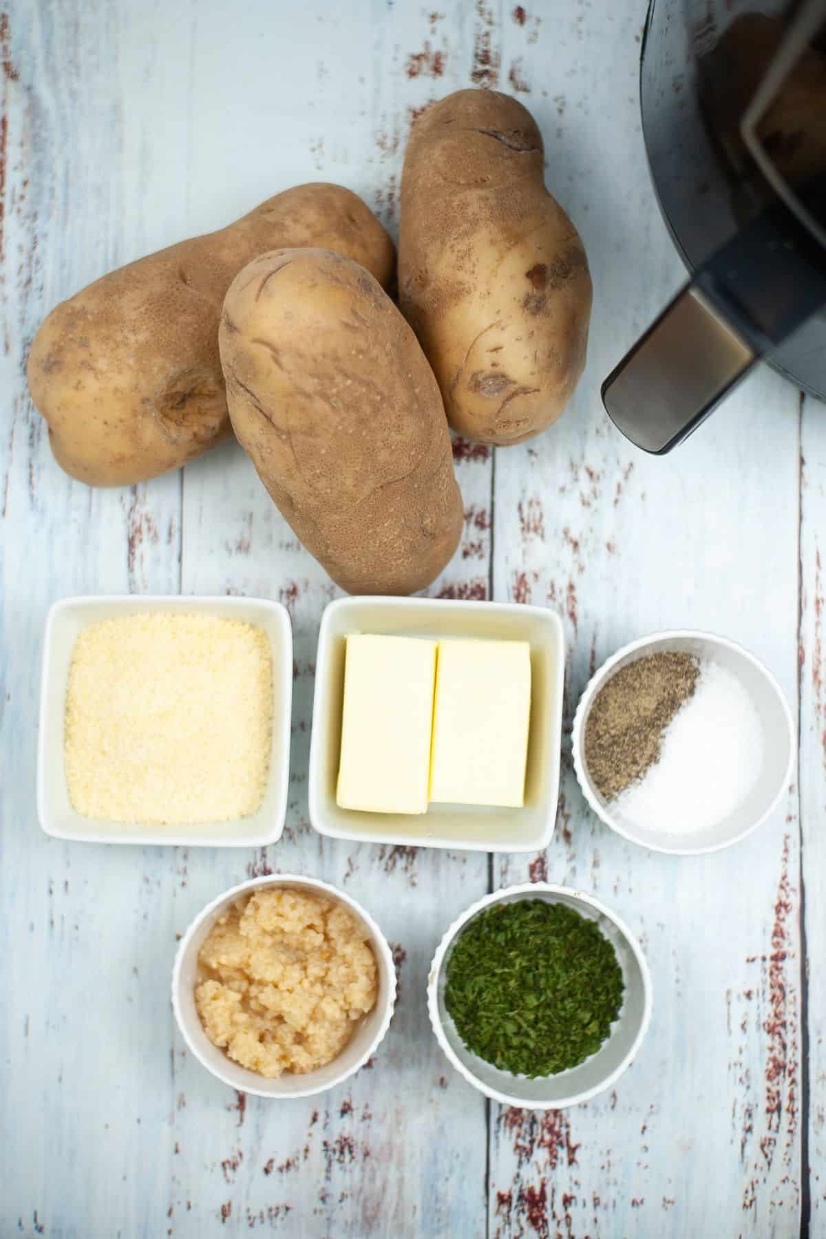 Three russet potatoes,with bowls of grated cheese, butter, salt, pepper, minced garlic, and chopped herbs on a rustic white wooden surface.