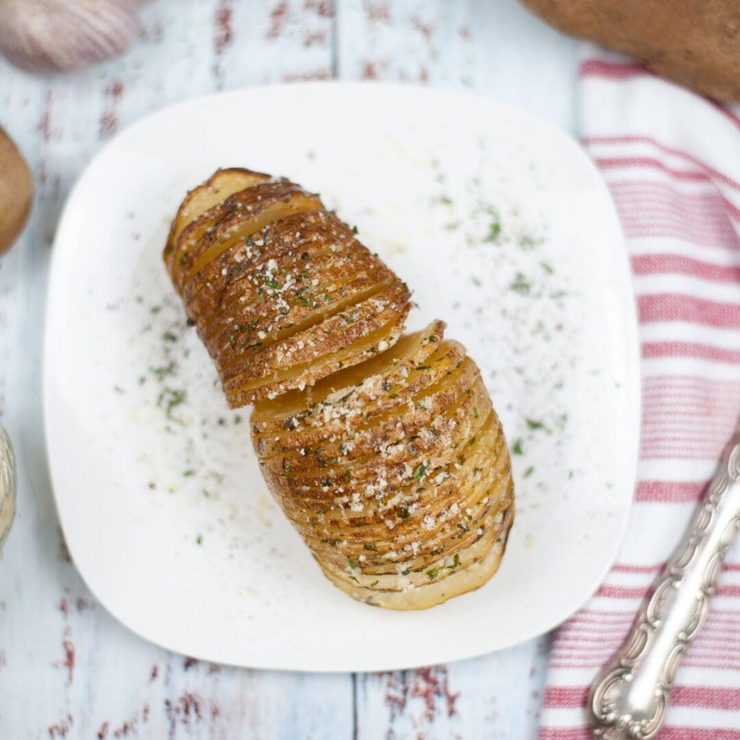 Air Fryer Hasselback Potatoes—sliced and roasted to perfection—are garnished with herbs and grated cheese, then served on a white plate beside a red and white striped towel.