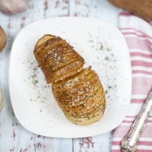 Air Fryer Hasselback Potatoes—sliced and roasted to perfection—are garnished with herbs and grated cheese, then served on a white plate beside a red and white striped towel.