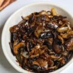A white bowl filled with savory mushrooms and onions, sautéed to perfection and caramelized, set on a white surface with a striped cloth in the background.