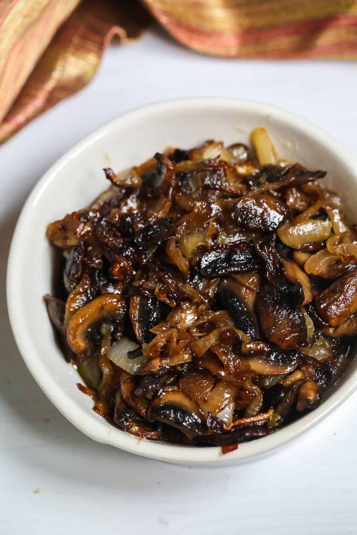 A white bowl filled with savory mushrooms and onions, sautéed to perfection and caramelized, set on a white surface with a striped cloth in the background.
