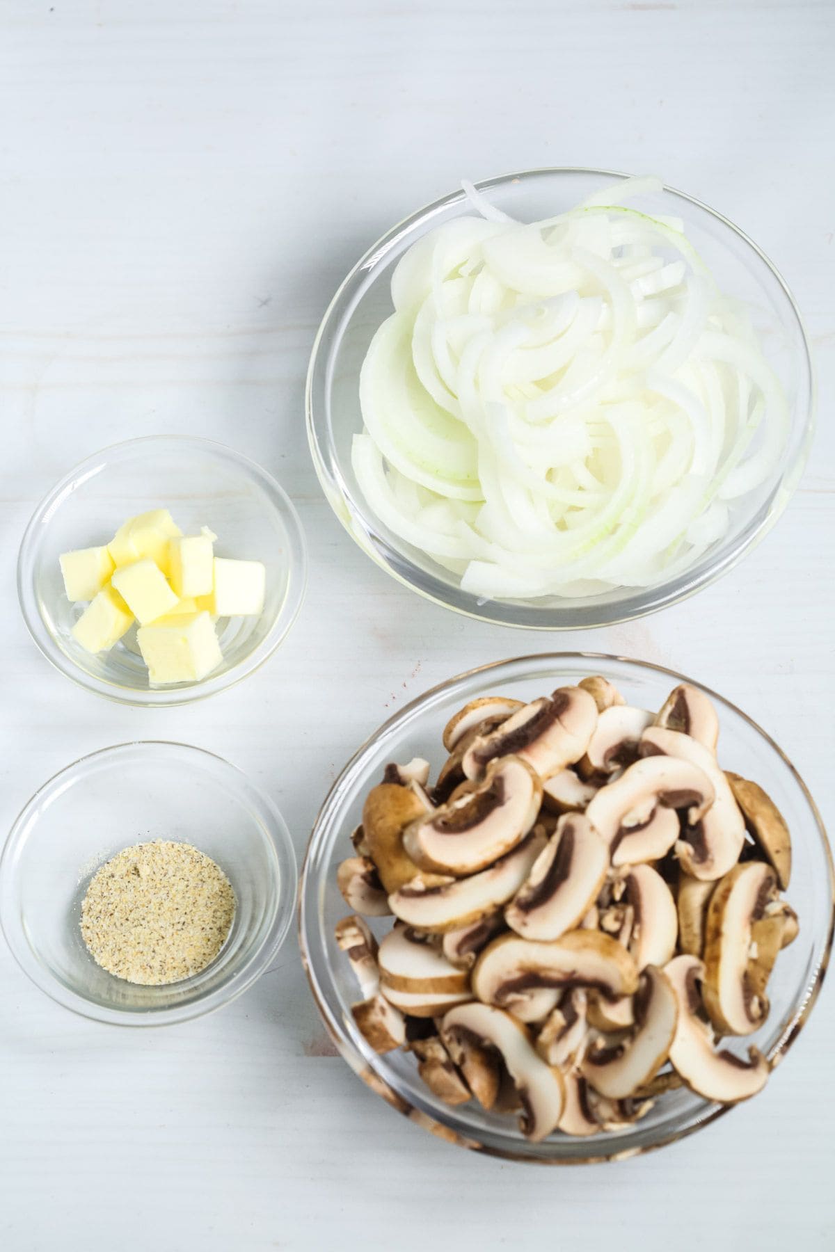 Four glass bowls containing sliced mushrooms and onions, cubed butter, and ground black pepper are arranged on a white surface.