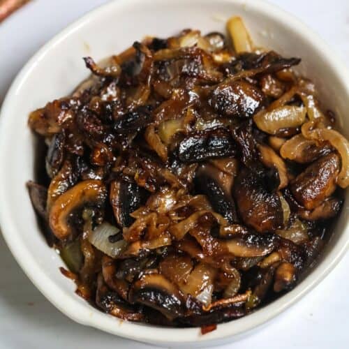 A white bowl filled with savory mushrooms and onions sits on a white surface, with caramelized flavors complemented by a striped cloth partially visible in the background.