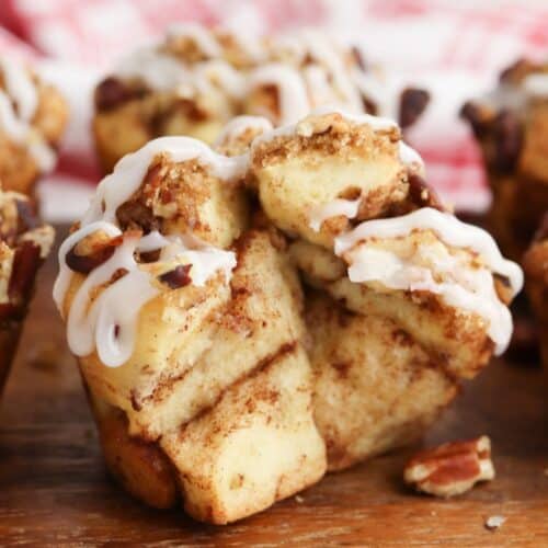 A close-up of Cinnamon Roll Muffins topped with white icing and small pieces of pecans, placed on a wooden surface.