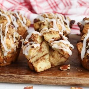 Close-up of Cinnamon Roll Muffins with icing and chopped nuts on top, arranged on a wooden board with a red-and-white cloth in the background.