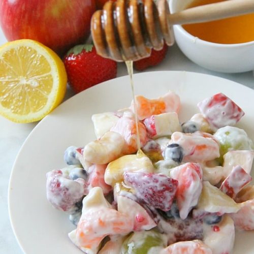 A bowl of fruit salad covered in a creamy dressing, being drizzled with honey