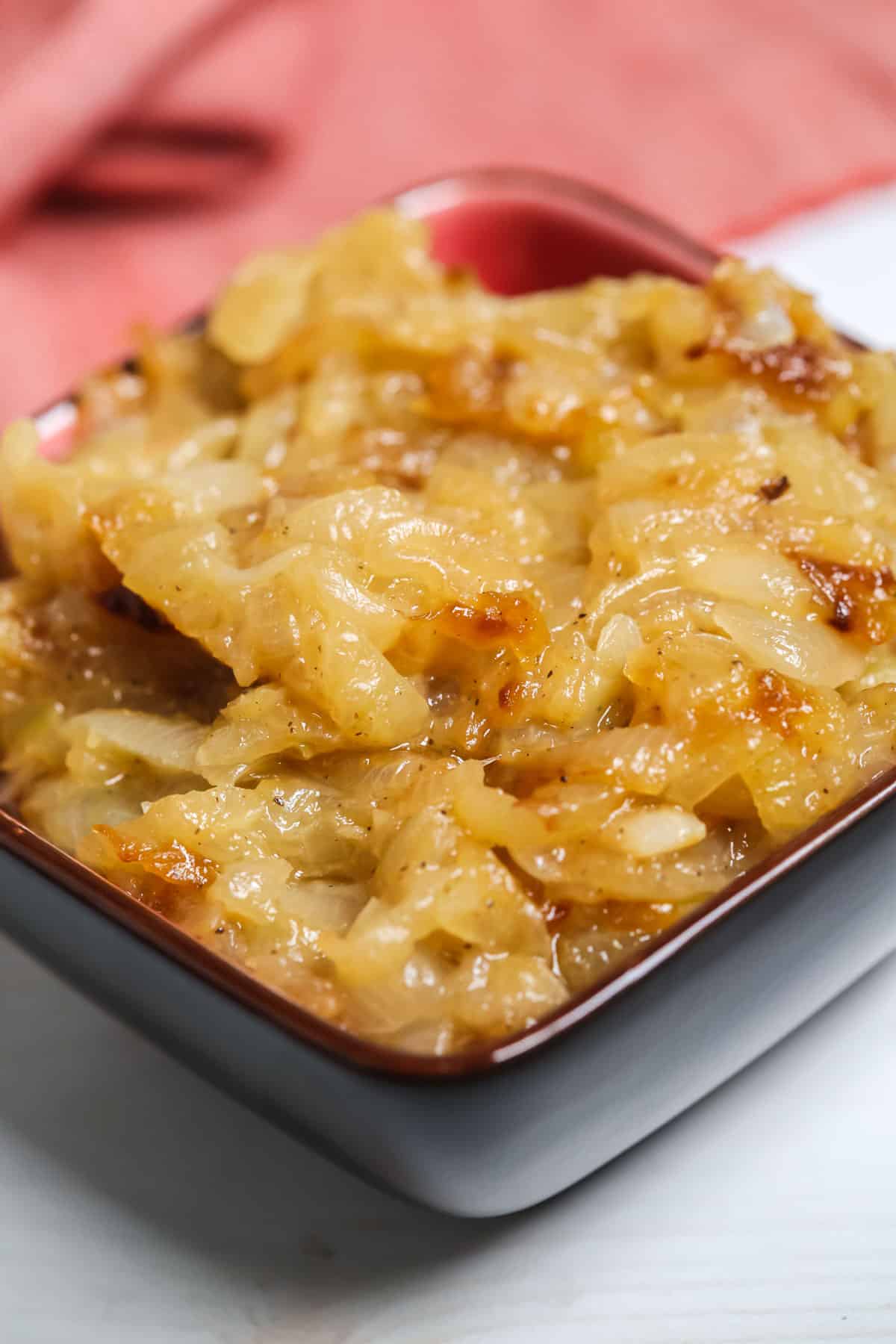 A close-up of Crockpot Caramelized Onions in a small square dish with a red interior on a white surface.
