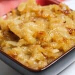 A square bowl filled with Crockpot Caramelized Onions displays a golden-brown, glossy texture. A red cloth is blurred in the background.