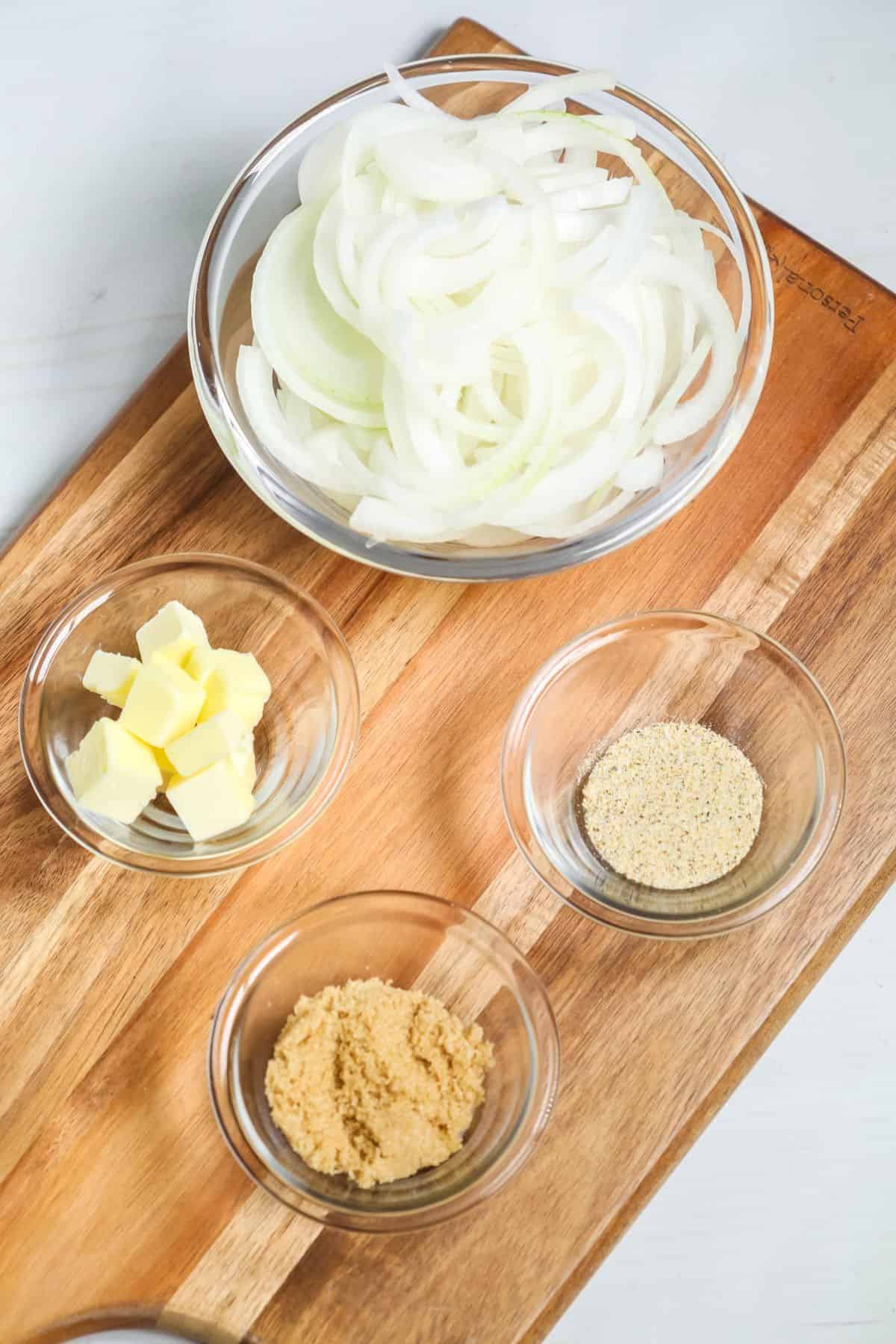 A bowl of sliced vegetables,butter cubes, brown sugar, and seasoning powder in separate glass bowls on a wooden cutting board.