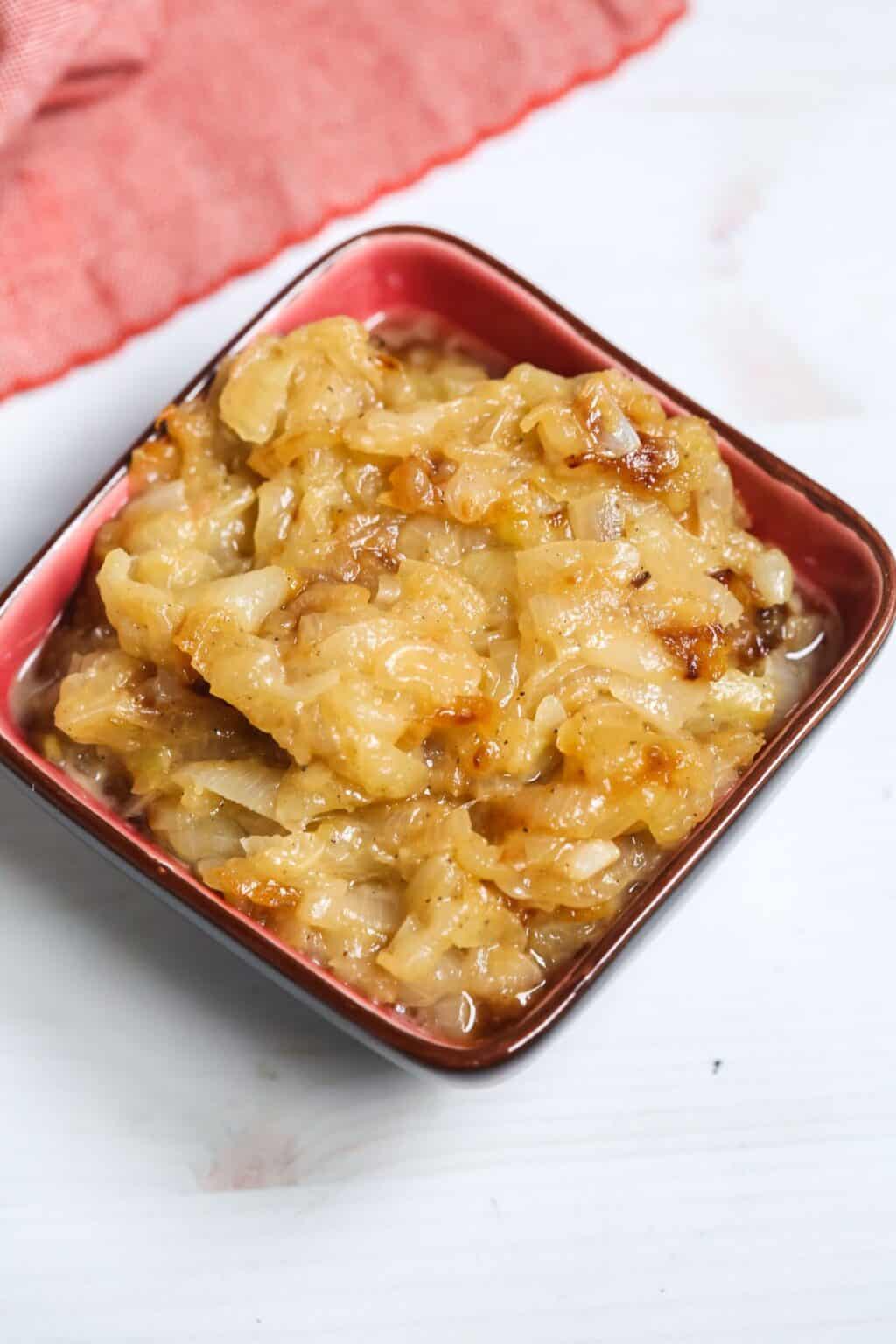 A small red bowl filled with Crockpot Caramelized Onions sits on a white surface, with a red cloth in the background.