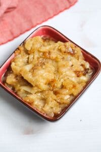A small red bowl filled with Crockpot Caramelized Onions sits on a white surface, with a red cloth in the background.