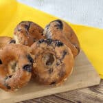 Five raisin bagels are displayed on a wooden cutting board, their golden hues contrasting beautifully with the yellow cloth background, reminiscent of freshly made glazed blueberry donuts.