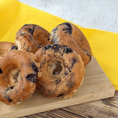 Five raisin bagels are displayed on a wooden cutting board, their golden hues contrasting beautifully with the yellow cloth background, reminiscent of freshly made glazed blueberry donuts.