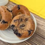 Three blueberry bagels on a white plate, placed on a wooden table with a yellow cloth in the background, evoke the charm of glazed blueberry donuts at a cozy morning brunch.
