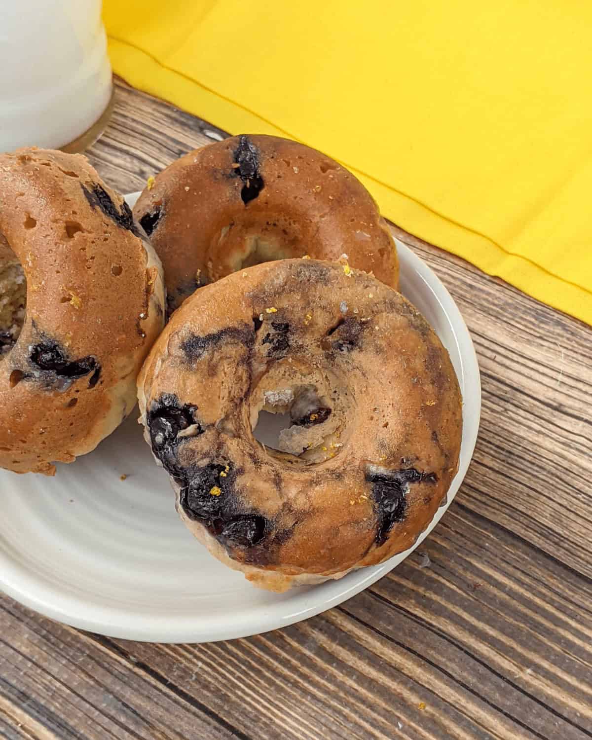 Three blueberry bagels on a white plate, placed on a wooden table with a yellow cloth in the background, evoke the charm of glazed blueberry donuts at a cozy morning brunch.