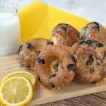 A stack of blueberry bagels, a glass of milk, and lemon slices are arranged on a wooden board with a yellow napkin in the background, reminiscent of glazed blueberry donuts.