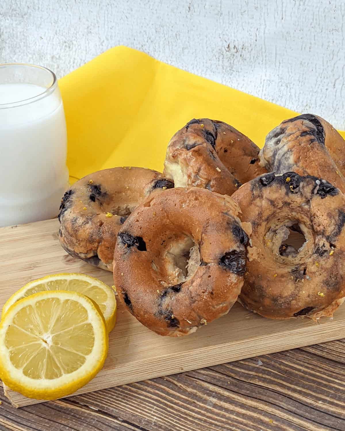 A stack of blueberry bagels, a glass of milk, and lemon slices are arranged on a wooden board with a yellow napkin in the background, reminiscent of glazed blueberry donuts.