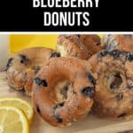 A wooden board holds several glazed blueberry donuts accompanied by a few lemon slices, with a small milk carton partially visible in the background.