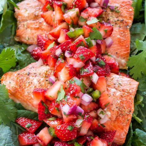 A closeup of several salmon filets on a bed of greens with strawberry jalepeno salsa on top