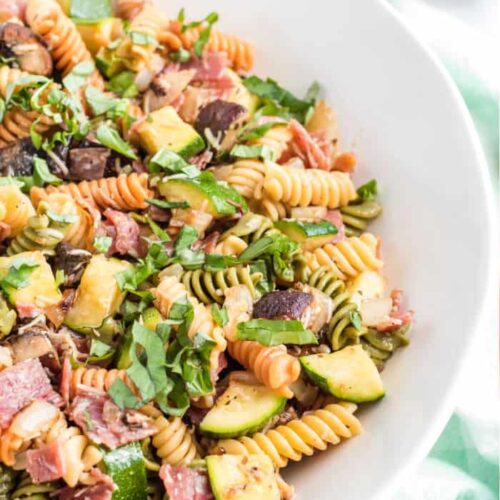A large bowl is filled with pasta, salami, and grilled vegetables