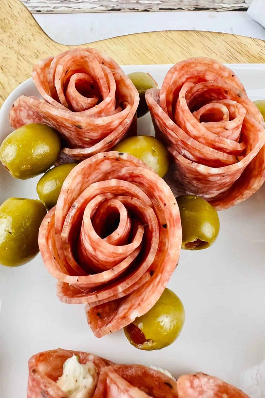 Slices of salami arranged in the shape of roses with green olives on a white plate, perfect for anyone learning how to make salami roses for an elegant appetizer.