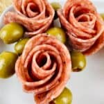 Slices of salami arranged in the shape of roses with green olives on a white plate, perfect for anyone learning how to make salami roses for an elegant appetizer.
