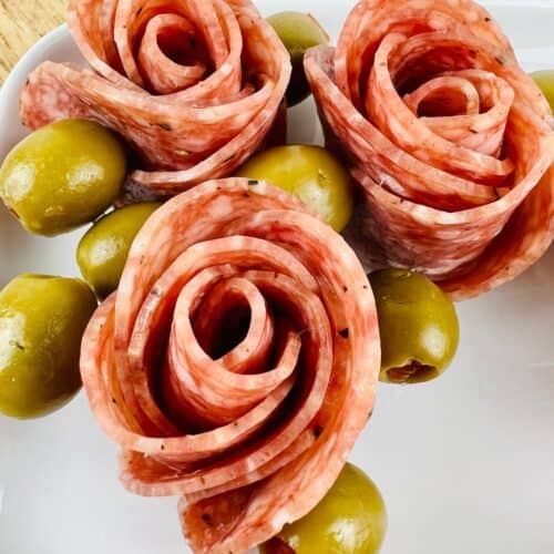 Slices of salami arranged in the shape of roses with green olives on a white plate, perfect for anyone learning how to make salami roses for an elegant appetizer.