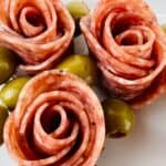 Slices of salami arranged in the shape of roses on a plate, each paired with green olives on toothpicks—learn how to make salami roses for an elegant charcuterie display.