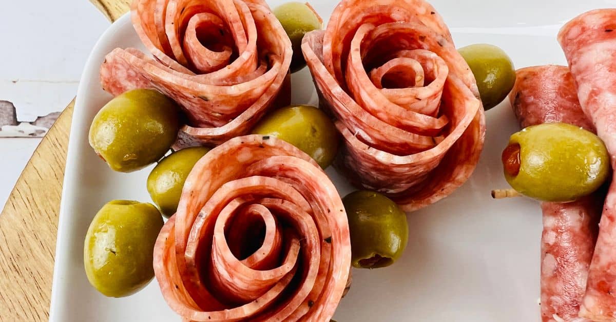 Slices of salami arranged in the shape of roses on a plate, each paired with green olives on toothpicks—learn how to make salami roses for an elegant charcuterie display.