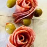 Slices of salami arranged in a rose shape, each garnished with three green olives, showcase how to make salami roses on a light-colored surface.
