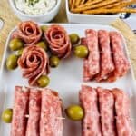A white platter with rolled salami slices, green olives, and beautiful salami roses—see How to Make Salami Roses—next to a bowl of dip with chives and a bowl of pretzel sticks.