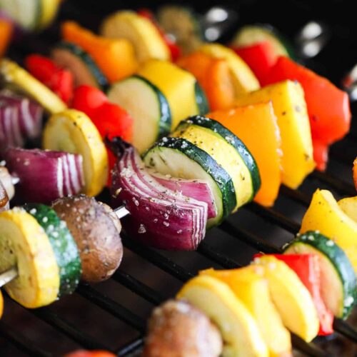 A grill has several colorful vegetable skewers featuring squash, zucchini, tomatoes, onions, and mushrooms