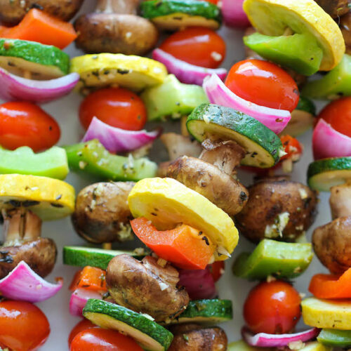 A plate of colorful vegetable kabobs featuring mushrooms, onions, squash, zucchini, and peppers