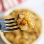 A close-up of a fork holding a bite of cheesy, baked macaroni with caramelized bits and Instant Pot Caramelized Onions, with a bowl of the same food in the background.