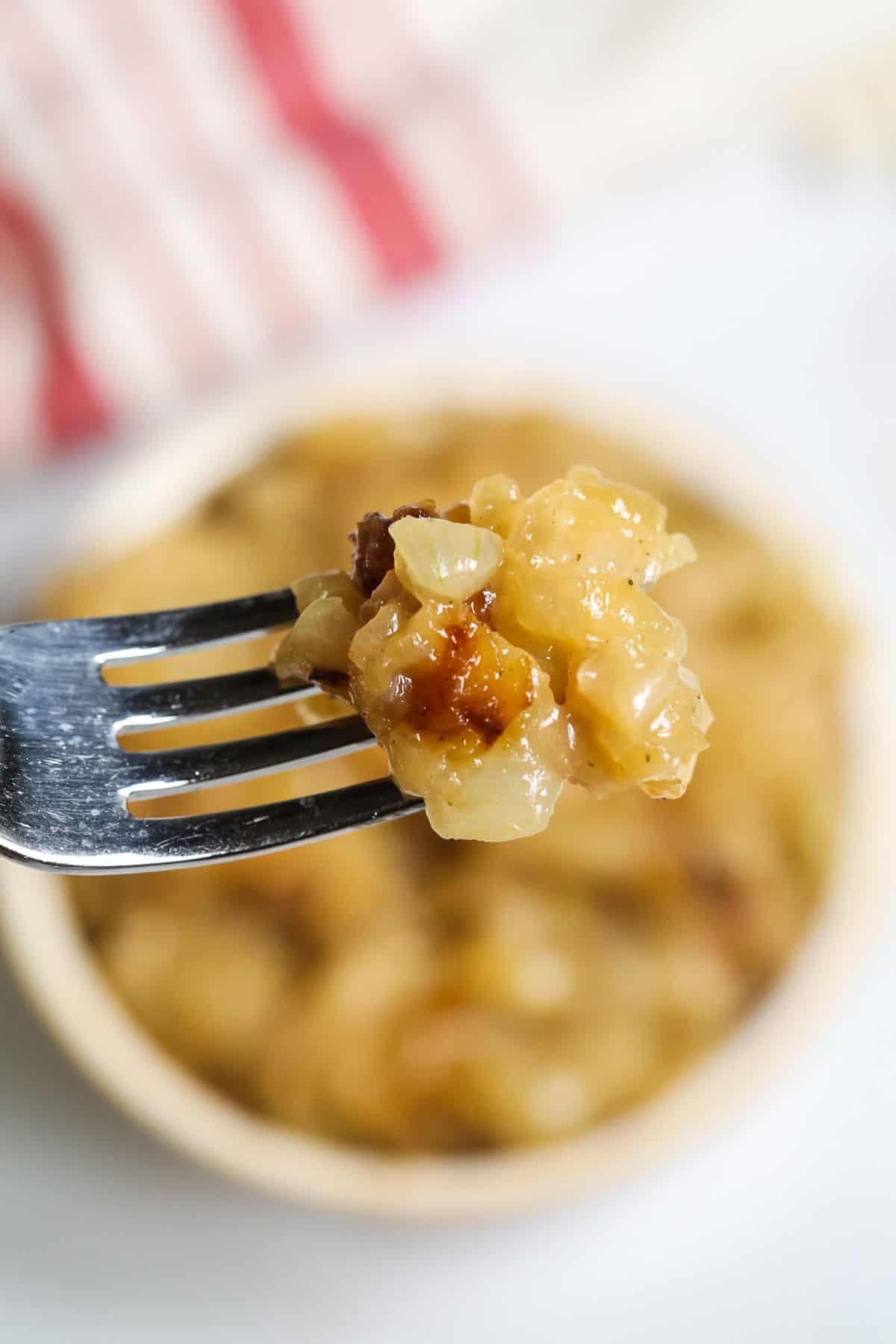 A close-up of a fork holding a bite of cheesy, baked macaroni with caramelized bits and Instant Pot Caramelized Onions, with a bowl of the same food in the background.