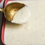 A spoon holding a portion of thick, creamy Tiger Sauce hovers above a square dish filled with the same zesty, white sauce.