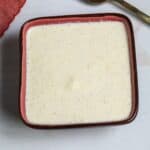 A square bowl mixture sits on a white surface next to a gold spoon and a red cloth.