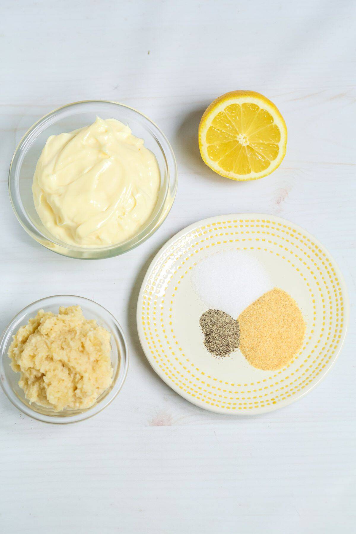 A bowl of mayonnaise, minced garlic, half a lemon, and a plate with salt, black pepper, sugar, and garlic powder are arranged on a white surface—perfect for crafting your own flavorful Tiger Sauce.
