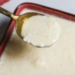 A spoon holding thick, creamy white Tiger Sauce with visible specks hovers above a bowl filled with the same zesty sauce.