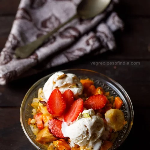 A small bowl of fruit salad with ice cream sits in front of a napkin with a spoon on it